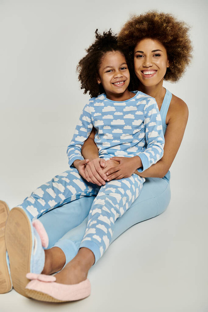 Feliz madre afroamericana y su hija sentadas en el suelo en pijamas azules a juego, compartiendo un momento acogedor juntos. - Foto, Imagen