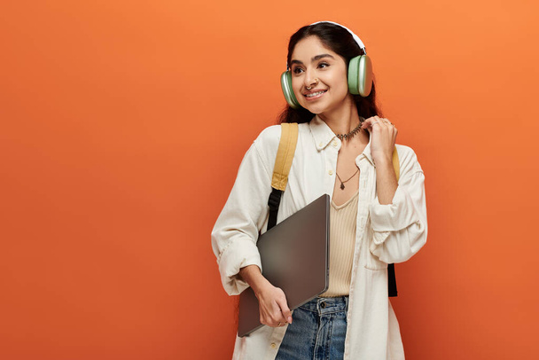 Kulaklıklı genç Hintli kadın, turuncu arka planda dizüstü bilgisayarı tutuyor.. - Fotoğraf, Görsel