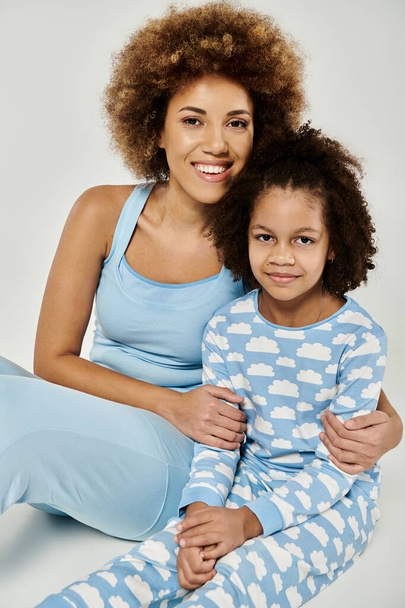 Un gioioso abbraccio tra madre e figlia afroamericana, in posa in pigiama blu abbinato su un morbido sfondo grigio. - Foto, immagini