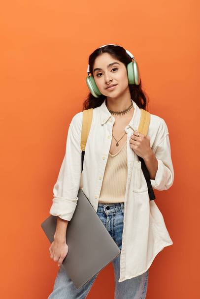 Genç Hintli kadın, turuncu duvara kulaklık takarken dizüstü bilgisayarını enerjik bir şekilde tutuyor.. - Fotoğraf, Görsel