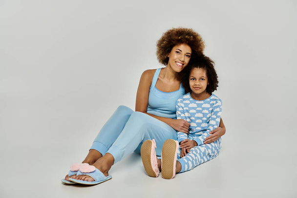Glückliche afroamerikanische Mutter und Tochter sitzen zusammen im Pyjama auf dem grauen Boden und teilen einen besonderen Moment. - Foto, Bild