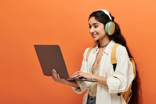 Jeune femme indienne avec écouteurs utilisant un ordinateur portable sur fond orange. - Photo, image