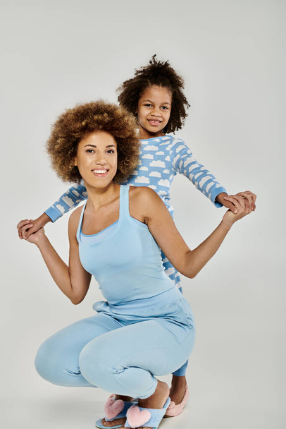 Gülümseyen Afro-Amerikan anne ve kızı aynı mavi pijamaları giyerek gri bir arka plana karşı birlikte poz veriyorlar.. - Fotoğraf, Görsel