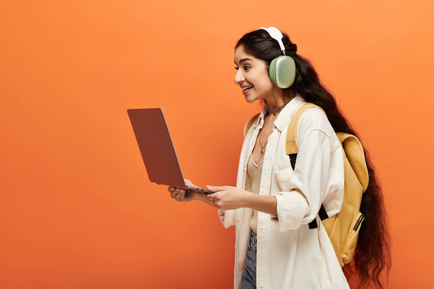 活気のあるオレンジ色の背景でラップトップを使用してヘッドフォンを持つ若いインドの女性. - 写真・画像