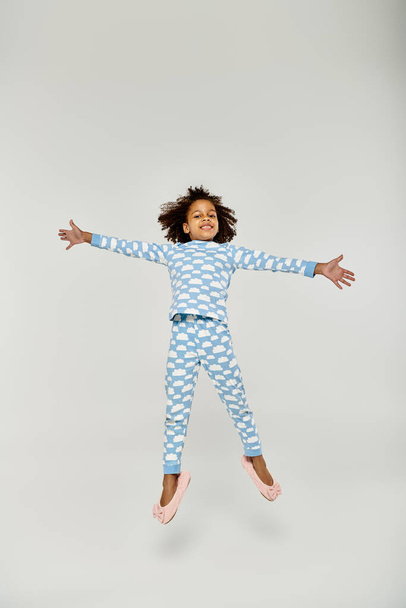Una chica afroamericana feliz saltando alegremente en un pijama azul con su madre cerca sobre un fondo gris. - Foto, imagen