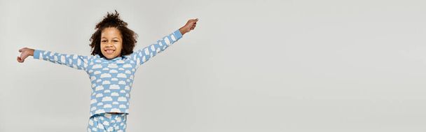Een vrolijk Afrikaans-Amerikaans meisje in pyjama dat haar armen uitstrekt over een grijze achtergrond. - Foto, afbeelding