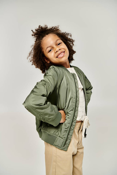 Кудрявая афроамериканка в зеленой куртке и коричневых штанах на сером фоне.. - Фото, изображение