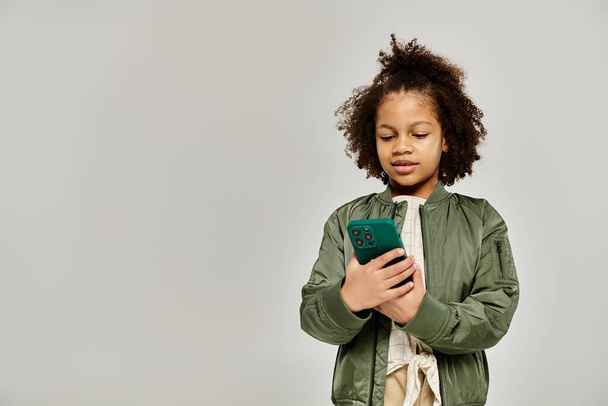 Une afro-américaine élégamment habillée aux cheveux bouclés tient un smartphone tout en se tenant debout sur un fond gris. - Photo, image