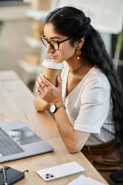 Eine professionelle indische Frau sitzt an einem Schreibtisch mit Laptop und Kaffeetasse. - Foto, Bild