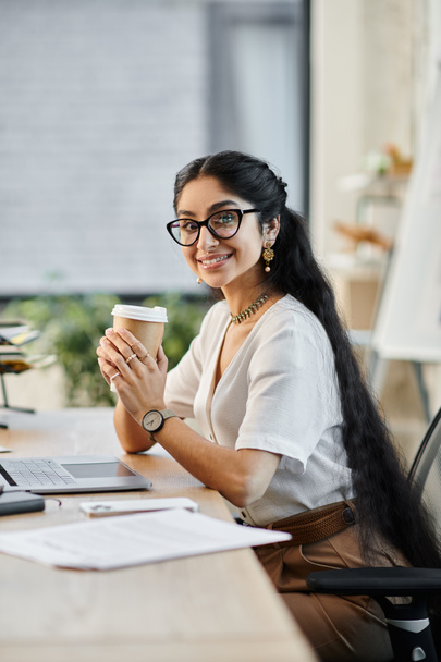 Een jonge indiaanse vrouw met een bril geniet van een kopje koffie aan haar bureau. - Foto, afbeelding