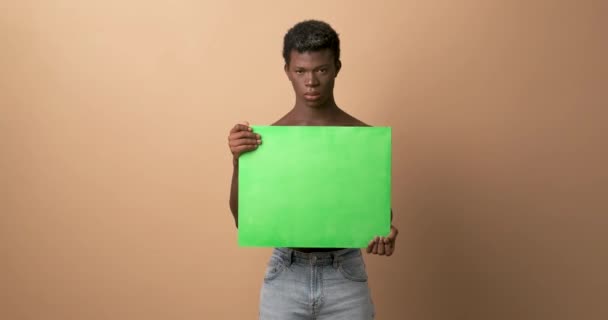 Yeşil renkli pankartı olan kızgın, genç, siyah bir adam stüdyoda bej arka planda kameraya bakıyor. - Video, Çekim