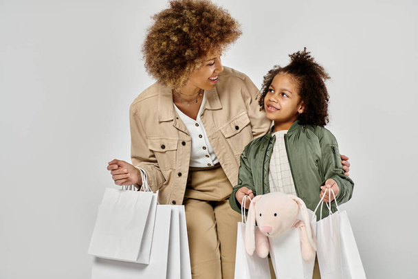 カーリーなアフリカ系アメリカ人の母親と娘は,グレーの背景に愛と喜びを示すショッピングバッグを持っています. - 写真・画像