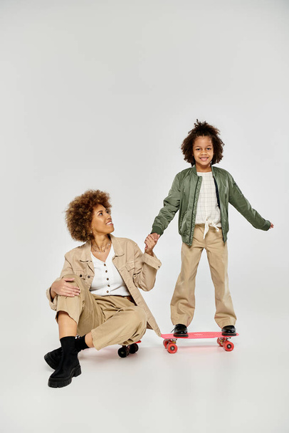 La madre y la hija afroamericanas rizadas con ropa elegante disfrutan de una divertida sesión de skate juntas sobre un fondo gris. - Foto, Imagen