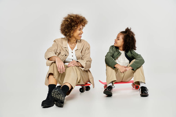 カーリー・アフリカ系アメリカ人の母親と娘は,灰色の背景に対してスケートボードの上にスタイリッシュに座ります.. - 写真・画像