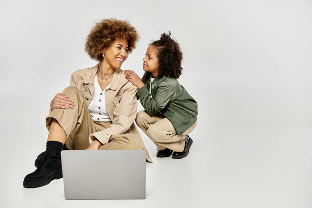 Σγουρή Αφρο-Αμερικανίδα μητέρα και κόρη με κομψά ρούχα, κάθονται στο πάτωμα, επικεντρώνονται σε μια οθόνη laptop. - Φωτογραφία, εικόνα