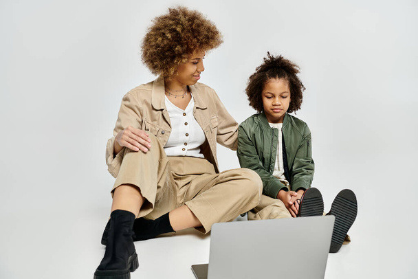Curly African American mother and daughter in stylish clothes sit on the floor, using a laptop together. - Photo, Image