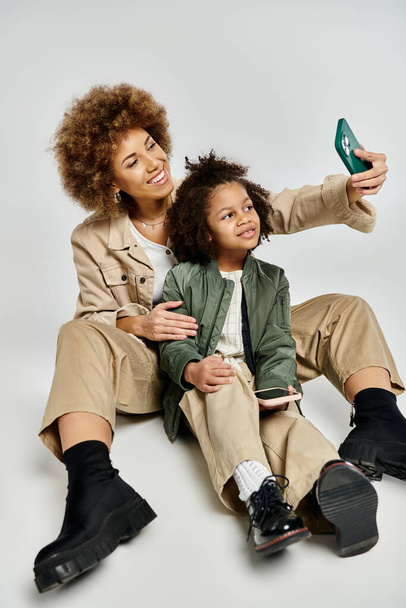 Кучерява афроамериканська мати і дочка в стильному одязі, приймаючи селфі з мобільним телефоном на сірому фоні. - Фото, зображення
