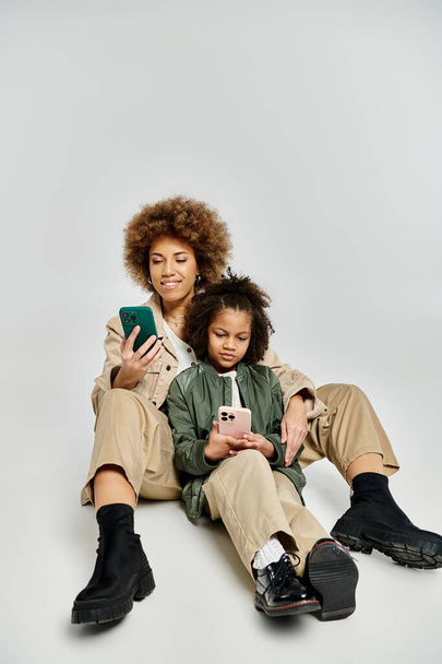 Μια σγουρή Αφρο-Αμερικανίδα μητέρα και κόρη με κομψά ρούχα που κάθονται στο πάτωμα, βαθιά απορροφημένοι από τη χρήση ενός κινητού τηλεφώνου. - Φωτογραφία, εικόνα