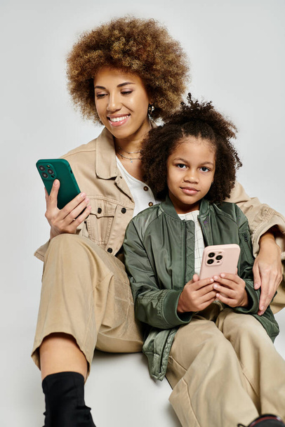 Madre e hija afroamericanas rizadas sentadas en el suelo, sosteniendo teléfonos celulares, con ropa elegante sobre fondo gris. - Foto, imagen