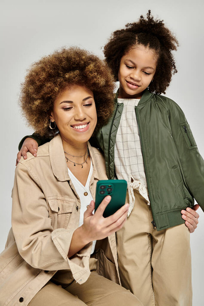 Kędzierzawa Afroamerykańska matka i córka, stylowo ubrana, pochłonięta telefonem komórkowym na szarym tle. - Zdjęcie, obraz
