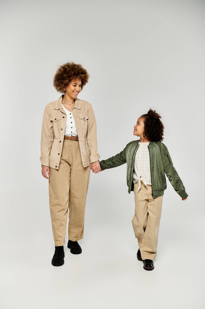 Curly afro-americano mãe e filha de mãos dadas em roupas elegantes contra um fundo de estúdio cinza. - Foto, Imagem