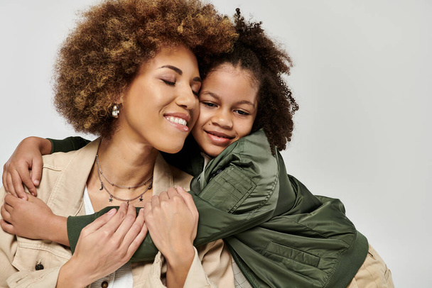 Кудрявые афроамериканская мать и дочь в стильной одежде обнимаются яростно на белом фоне. - Фото, изображение
