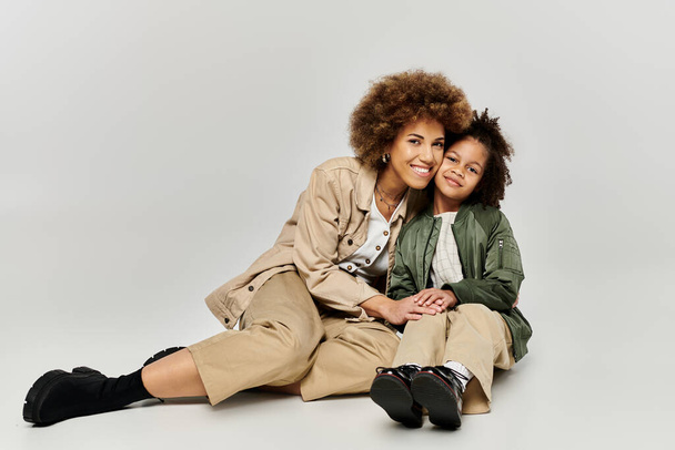 krullend Afro-Amerikaanse moeder en dochter in stijlvolle kleding, zitten op de vloer, glimlachen warm naar elkaar. - Foto, afbeelding