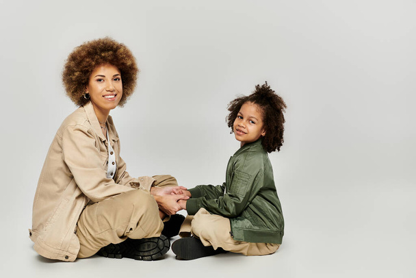 Σγουρή Αφρο-Αμερικανίδα μητέρα και κόρη κάθονται μαζί στο γκρι πάτωμα με κομψά ρούχα, μοιράζονται μια τρυφερή στιγμή. - Φωτογραφία, εικόνα