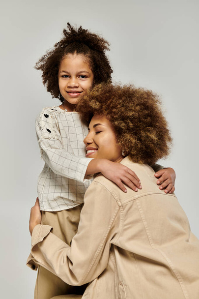 Göndör afro-amerikai anya és lánya elegáns ruhákban egy szívmelengető ölelés egy szürke háttér. - Fotó, kép