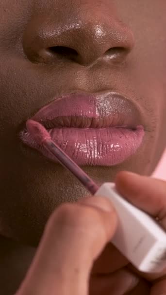 Primer plano de la cosecha visagiste irreconocible aplicando brillo de labios con cepillo mientras se hace el maquillaje en drag queen afroamericano con una piel lisa perfecta sobre fondo rosa. Imágenes verticales - Metraje, vídeo