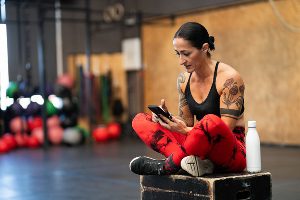 Sportowa dojrzała kobieta korzystająca z telefonu komórkowego zrelaksowana na treningu crossowym - Zdjęcie, obraz