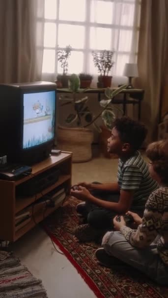 Pystysuora puoli handheld kuvamateriaalia kaksi monipuolista poikaa istuu lattialla edessä putki-TV ja pelaa vanhan koulukunnan arcade videopeli retro-tyylistä kotona, 90-luku - Materiaali, video