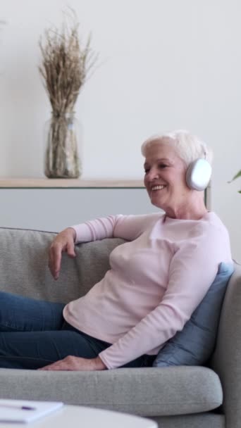 Starší šťastná běloška, která poslouchá hudbu v bezdrátových sluchátkách a odpočívá doma na pohovce. Rekreace, potěšení, koncept občerstvení. Svislé video. - Záběry, video