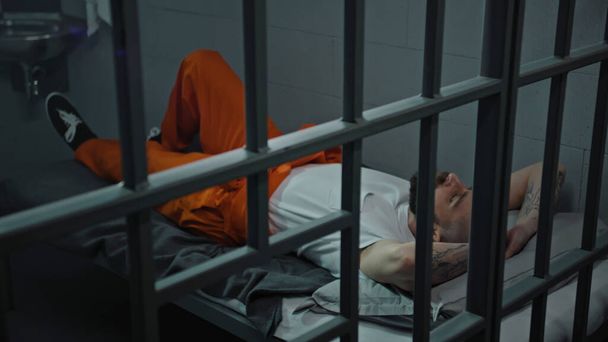 Gevangene in oranje uniform ligt op het celbed. Gevangene zit gevangenisstraf uit voor misdaad. Crimineel in detentiecentrum, gevangenis. Justitie. Zicht door metalen staven. Dolly schot. - Foto, afbeelding