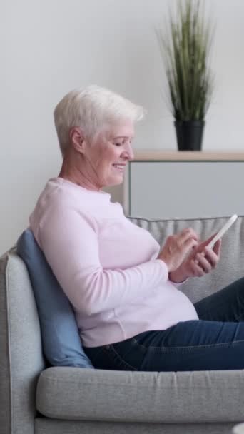 Idősebb kaukázusi nő, aki mobilportréval böngészi az internetet, és otthon a kanapén nézi a kamerát. Pihenés, pihenés. Függőleges videó. - Felvétel, videó