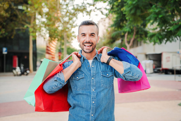 Vain yksi nuori aikuinen kaveri kantaa ostoskasseja ostettuaan vaatteita ostoskeskuksesta. Todellinen onnellinen mies asiakas näyttää paketteja, jotka hän ostaa musta perjantai alennus. Shopaholainen mies hymyilee - Valokuva, kuva