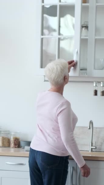 Iäkäs onnellinen valkoihoinen nainen kattamassa ruokapöytää kotona keittiössä. Tarjoilu astioiden kanssa ja juhla-aterian valmistelu. Pystysuora video. - Materiaali, video
