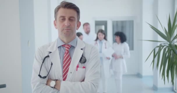 Confident, male specialist posing with folded arms at the clinic hall. Doctors working in the background. Copy space. - Footage, Video