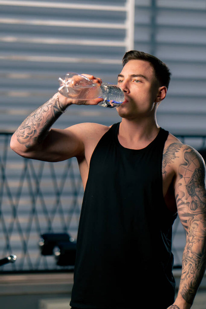 en el gimnasio un chico guapo joven con tatuajes en una camiseta negra bebe agua de una botella - Foto, imagen