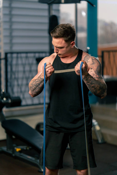 в спортзале молодой красивый парень с татуировками в черной футболке делает упражнения с эластичной лентой - Фото, изображение