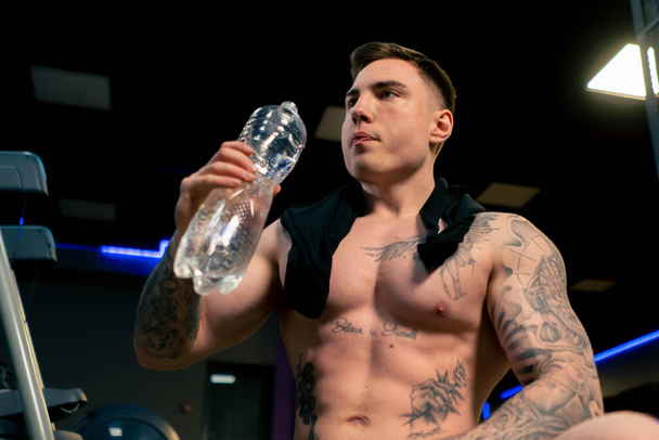κοντά στο γυμναστήριο νεαρός όμορφος άντρας με τατουάζ κάθεται πόσιμο νερό από ένα πλαστικό μπουκάλι ανάπαυσης - Φωτογραφία, εικόνα
