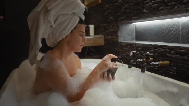 Een serene vrouw met een handdoek op haar hoofd ontspant in een bubbelbad, met behulp van zeep binnen. - Video