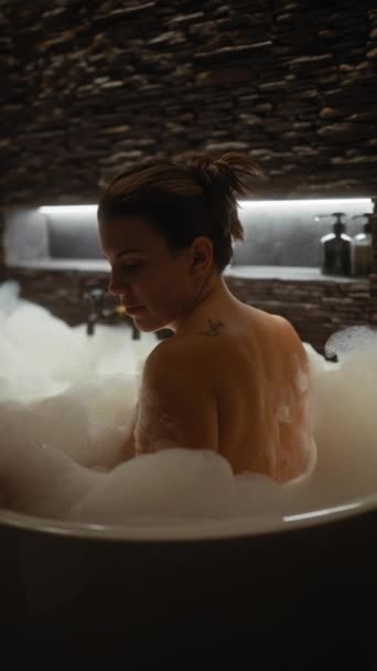 Una mujer serena disfruta de un baño de burbujas en un acogedor entorno de baño de paredes de piedra, que exuda tranquilidad y cuidado personal. - Metraje, vídeo