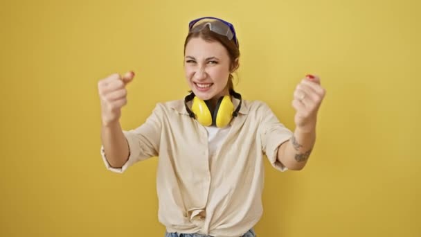 Excited and proud! young brunette girl celebrates joyous victory, arms raised, rocking safety glasses and noise reduction headphones against a bright yellow isolated background. - Footage, Video