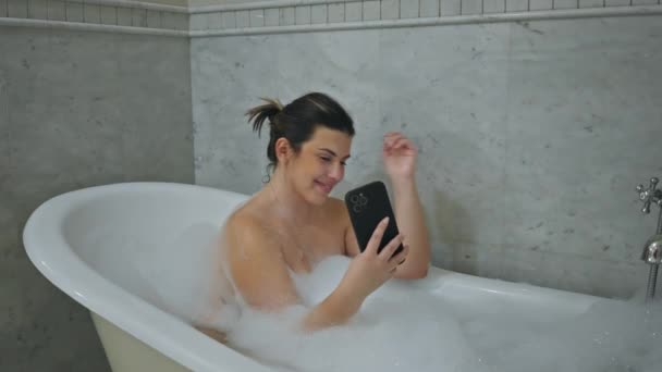 Una donna rilassata gode di un bagno di bolle mentre utilizza uno smartphone in un bagno moderno. - Filmati, video