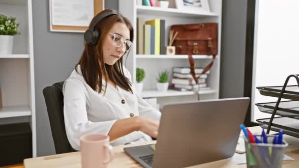 A young brunette woman joyfully dancing in an office setting, listening to music with headphones while using a laptop. - Footage, Video