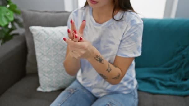 Uma jovem examina sua dor de mão dentro de casa, sentada em um sofá com uma tatuagem visível. - Filmagem, Vídeo