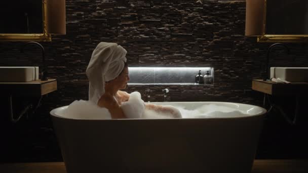 Huzurlu bir kadın, yumuşak ışıkla aydınlatılmış modern bir banyoda lüks bir köpük banyosunun keyfini çıkarıyor.. - Video, Çekim