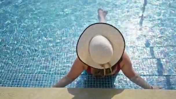 A woman lounges at a resort's swimming pool wearing a wide-brimmed hat, evoking a serene summer vibe. - Footage, Video