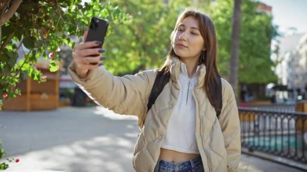 Een jonge vrouw neemt een selfie met een smartphone in een zonnig stadspark. - Video